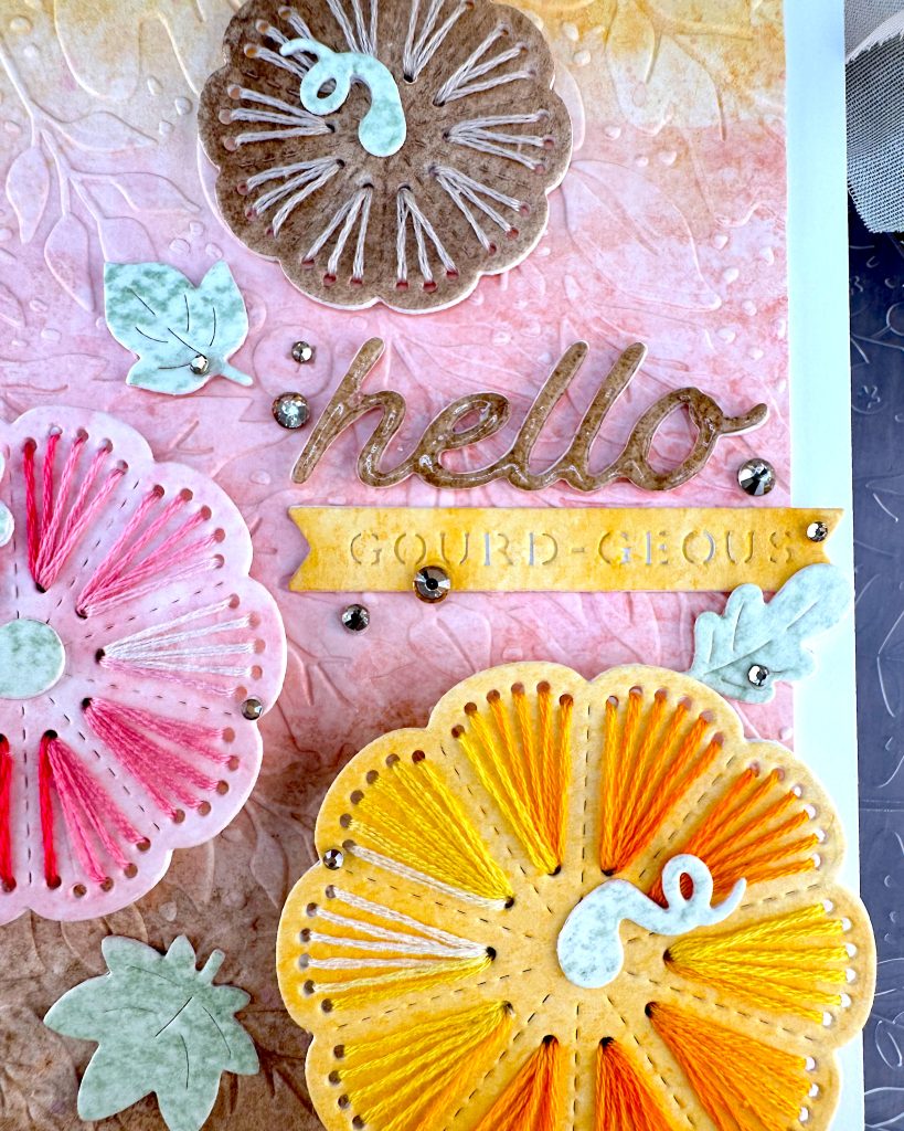 A handmade greeting card with a stitched pumpkin design sits on a wooden surface surrounded by craft supplies. The card features a textured gradient background that transitions from yellow at the top through coral-pink in the middle to brown at the bottom.
Three large pumpkins stitched with embroidery floss are prominently displayed: a small brown pumpkin embroidered with tan floss at the top, a pink pumpkin stitched with variegated red floss on the left, and a large, bright yellow pumpkin stitched with variegated yellow floss at the bottom right.
The word "hello" appears in glossy brown cursive lettering in the center, with a small yellow banner below it. The banner reads "GOURD-EOUS." Small mint green leaf embellishments and gold rhinestone accents are scattered throughout the design.
Under the card an embossing folder with a leaf pattern and a few pumpkin stitching dies are visible. So are some pale pink fabric leaves and mini pumpkins.
