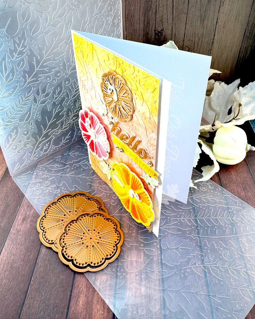 A handmade greeting card with a stitched pumpkin design sits on a wooden surface surrounded by craft supplies. The card features a textured gradient background that transitions from yellow at the top through coral-pink in the middle to brown at the bottom.
Three large pumpkins stitched with embroidery floss are prominently displayed: a small brown pumpkin embroidered with tan floss at the top, a pink pumpkin stitched with variegated red floss on the left, and a large, bright yellow pumpkin stitched with variegated yellow floss at the bottom right.
The photo is taken from the top right, allowing you to appreciate that the pink and yellow pumpkins are popped up on foam tape.
The word "hello" appears in glossy brown cursive lettering in the center, with a small yellow banner below it. The banner reads "GOURD-EOUS." Small mint green leaf embellishments and gold rhinestone accents are scattered throughout the design.
Under the card an embossing folder with a leaf pattern and a few pumpkin stitching dies are visible. So are some pale pink fabric leaves and mini pumpkins.
