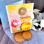 A handmade greeting card with a stitched pumpkin design sits on a wooden surface surrounded by craft supplies. The card features a textured gradient background that transitions from yellow at the top through coral-pink in the middle to brown at the bottom. Three large pumpkins stitched with embroidery floss are prominently displayed: a small brown pumpkin embroidered with tan floss at the top, a pink pumpkin stitched with variegated red floss on the left, and a large, bright yellow pumpkin stitched with variegated yellow floss at the bottom right. The word "hello" appears in glossy brown cursive lettering in the center, with a small yellow banner below it. The banner reads "GOURD-EOUS." Small mint green leaf embellishments and gold rhinestone accents are scattered throughout the design. Under the card an embossing folder with a leaf pattern and a few pumpkin stitching dies are visible. So are some pale pink fabric leaves and mini pumpkins.