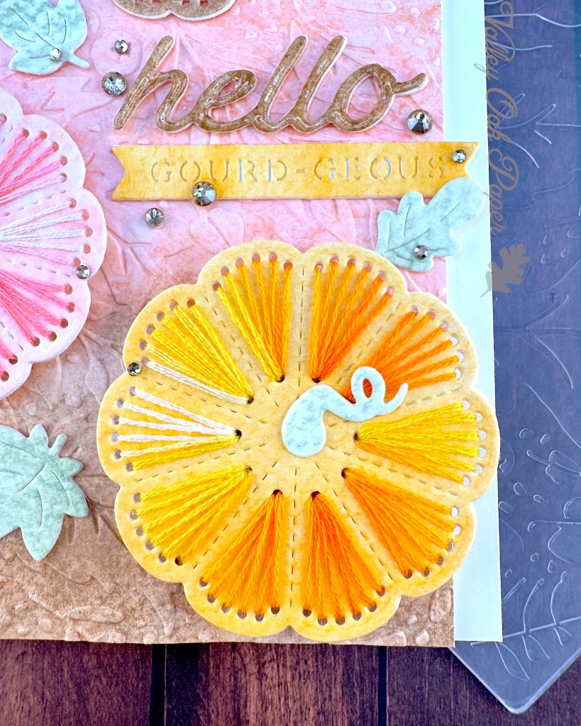 A handmade greeting card with a stitched pumpkin design sits on a wooden surface surrounded by craft supplies. The card features a textured gradient background that transitions from yellow at the top through coral-pink in the middle to brown at the bottom.
This is a closeup of the largest pumpkin and the sentiment. The HELLO is brown and covered with a clear, glossy lacquer. The GOURD-GEOUS is embossed into a yellow piece of watercolor paper with fish tails on the left and right ends.
The photo is taken from the top right, allowing you to appreciate that the pink and yellow pumpkins are popped up on foam tape.
The word "hello" appears in glossy brown cursive lettering in the center, with a small yellow banner below it. The banner reads "GOURD-EOUS." Small mint green leaf embellishments and gold rhinestone accents are scattered throughout the design.
Under the card an embossing folder with a leaf pattern and a few pumpkin stitching dies are visible. So are some pale pink fabric leaves and mini pumpkins.