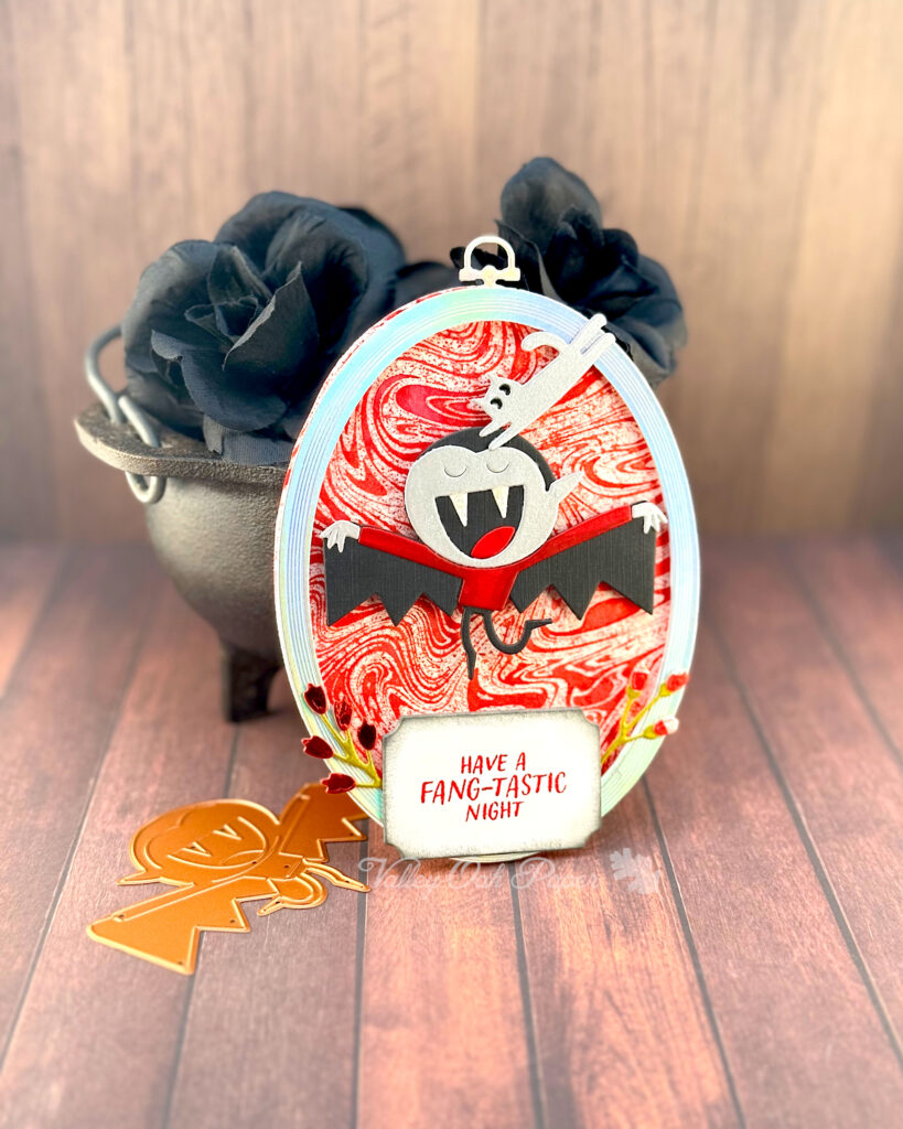 An oval greeting card with a blood red marbled background leans against a mini cauldron filled with black roses. 

The main feature of the card is a vampire wearing a black cape. A silver cat is jumping from the frame onto his head. 

The greeting reads "Have a Fang-Tastic Night."