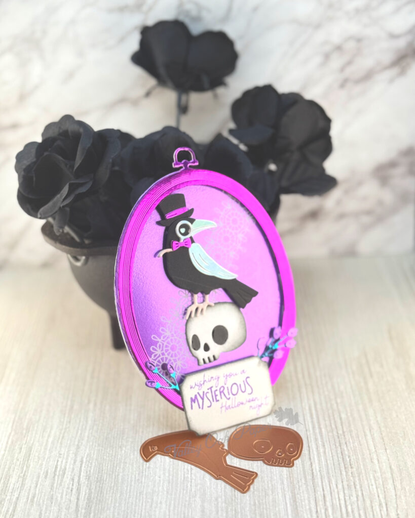 An oval greeting card leans against a mini cauldron filled with black roses. 

The main feature of the card is a black raven wearing a purple bow tie, a top hat and silver monocle. The raven is standing on a human skull.

The background is purple with a lace pattern. The greeting reads "Wishing you a mysterious Halloween night."