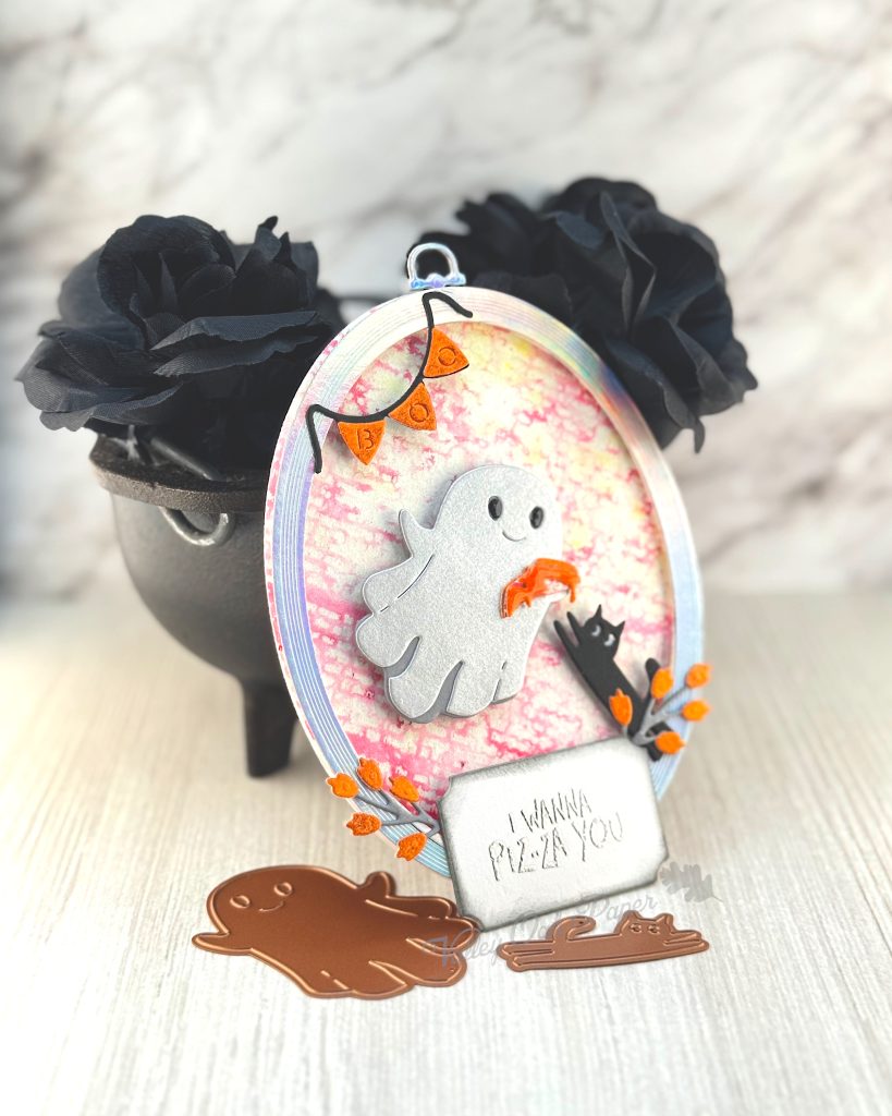 An oval greeting card leans against a mini cauldron filled with black roses. 

The main feature of the card is a silvery ghost holding a slice of pizza. A black cat is reaching up toward the pizza. 

The background is red and white with small text. The greeting reads "I wanna a Piz-Za of You."