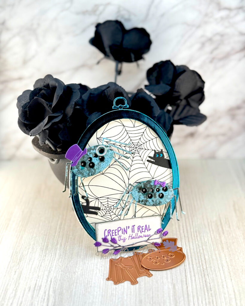 An oval greeting card, with a spider web background, leans against a mini cauldron filled with black roses. 

The card features two mottle green spiders with many eyes. One spider is wearing a purple top hat and monocle. The other one a purple hair tie. One black cat is racing into the frame, while another one appears to be racing out of the frame.

The greeting reads "Creepin' It Real this Halloween."