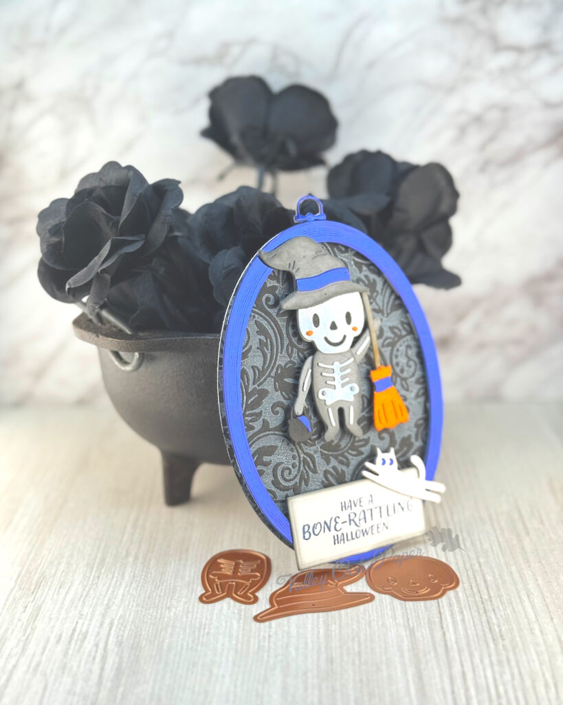 An oval greeting card, with a black damask pattern, leans against a mini cauldron filled with black roses. 

The main feature of the card is a little trick-or-treater. The child is dressed in a skeleton suit and a witch's hat. A white cat is running toward the little cauldron the child is holding in one hand. 

The greeting reads "Have a bone-rattling Halloween."