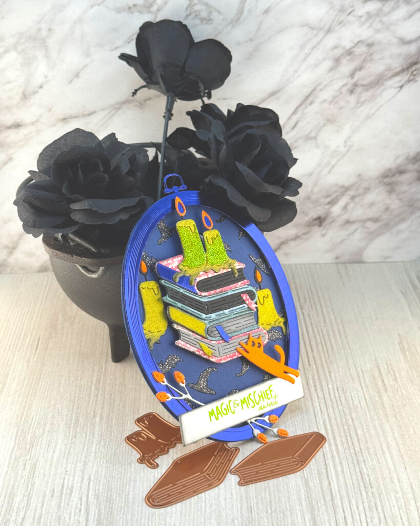 An oval greeting card with a dark blue background leans against a mini cauldron filled with black roses. 

The main feature of the card is a stack of hardcover books. They are surrounded by candles of a sickly green color. An orange cat is reaching up toward the ribbon bookmark sticking out from one of the books. 

The greeting reads "Magic & Mischief Awaits."