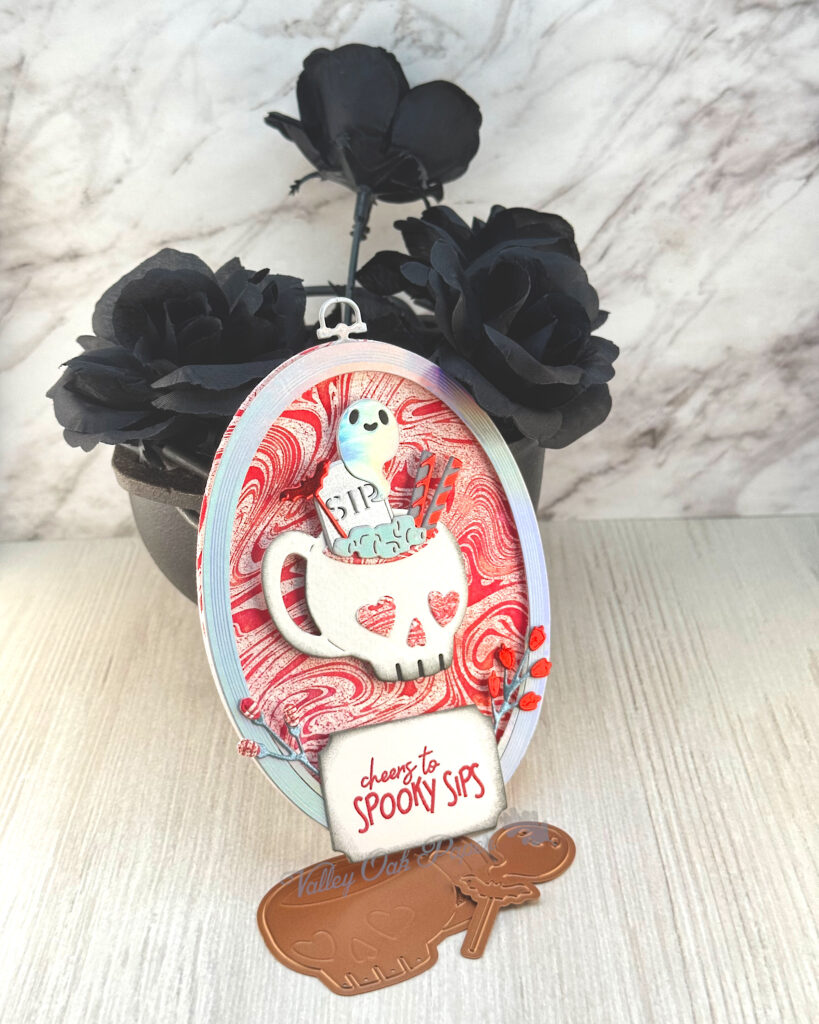 An oval greeting card, with a blood red marbled background, leans against a mini cauldron filled with black roses. 

The main feature of the card is a skull mug with a drink inside. The drink is decorated with a bat pick, a mini toombstone that reads "SIP" and straws. A silver ghost hovers above the drink.

The greeting reads "Cheers to Spooky Sips."