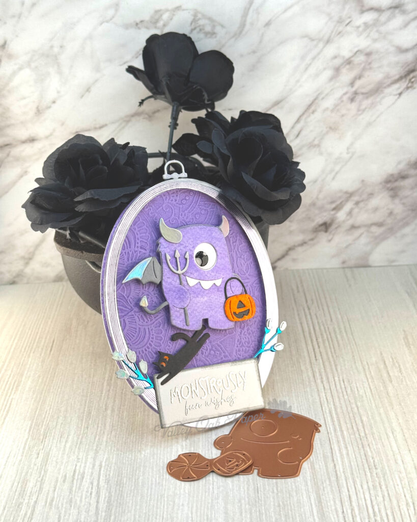 An oval greeting card with a purple background leans against a mini cauldron filled with black roses. 

The main feature of the card is a shaggy purple cyclop wielding a trident and a pumpkin bucket. A black cat is running away from the monster. 

The greeting reads "Monstrously Fun Wishes."