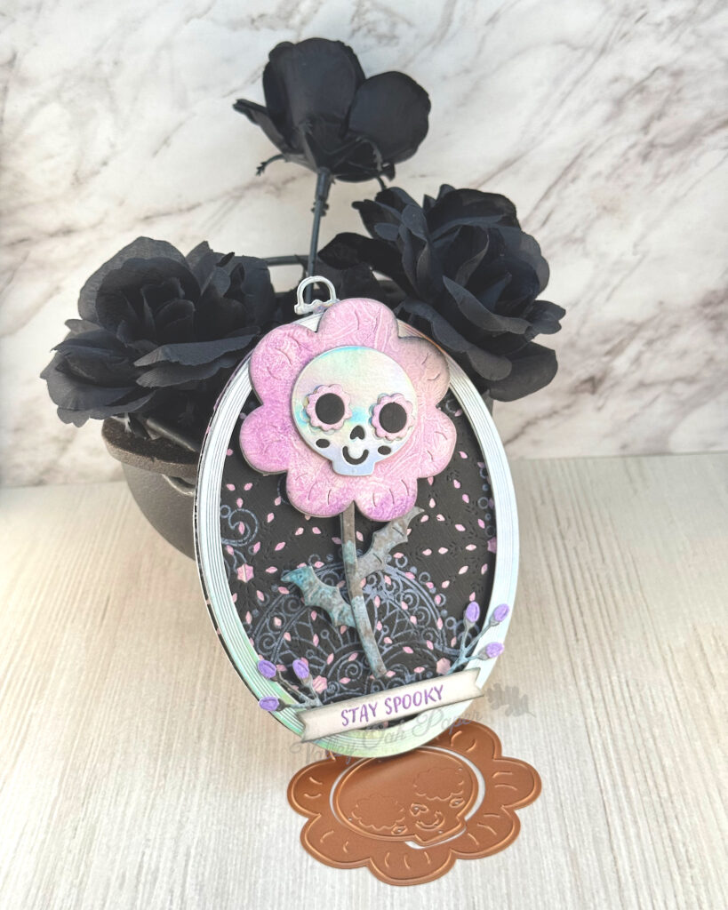An oval greeting card leans against a mini cauldron filled with black roses. 

The main feature of the card is an oversized pink flower with a skull in the flower center. The skull appears to be wearing pink flower-shaped sunglasses. 

The background is black with a pink pattern. The greeting reads "Stay Spooky."