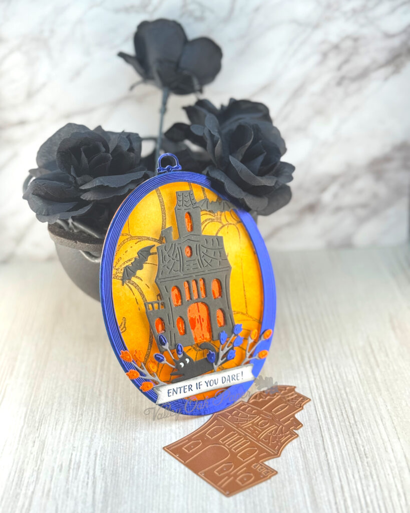 An oval greeting card leans against a mini cauldron filled with black roses. 

The main feature of the card is an old mansion with an eerie orange glow coming out of all the windows and doors. A black cat is skulking in the undergrowth in front of it.

The background is orange with a pumpkin pattern. The greeting reads "Enter If You Dare."