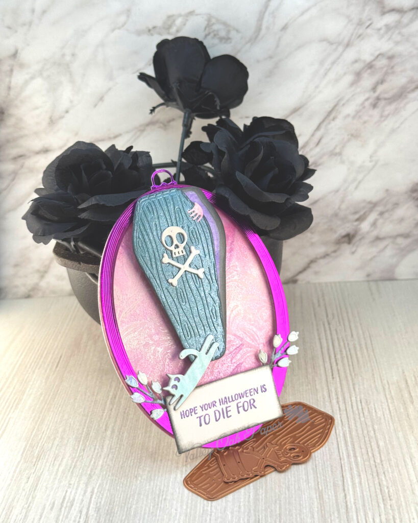 An oval greeting card with a pink background leans against a mini cauldron filled with black roses. 

The main feature of the card is a wooden casket. The lid is ajar and a hand is holding it open. A silver cat is jumping off the lid. 

The greeting reads "Hope Your Halloween is to Die For."