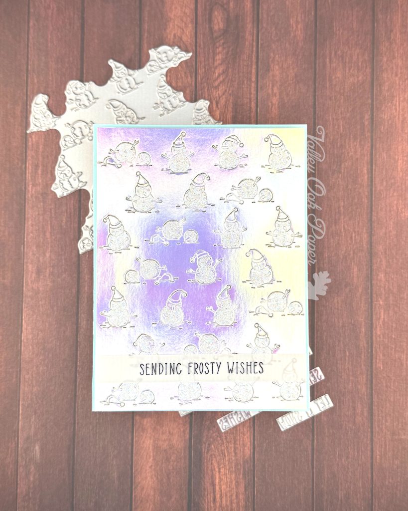 A handmade greeting card lies on top of a wooden surface. The background on the card is made of silver holographic cardstock. It's been embossed with a pattern of snow people. The snow is covered with iridescent glitter.
Toward the bottom of the card, a thin vellum strip reads "Sending Frosty Wishes."
A slim matte of pale blue is visible around the patterned holographic cardstock.
Peeking out from under the card are some BetterPress plates.