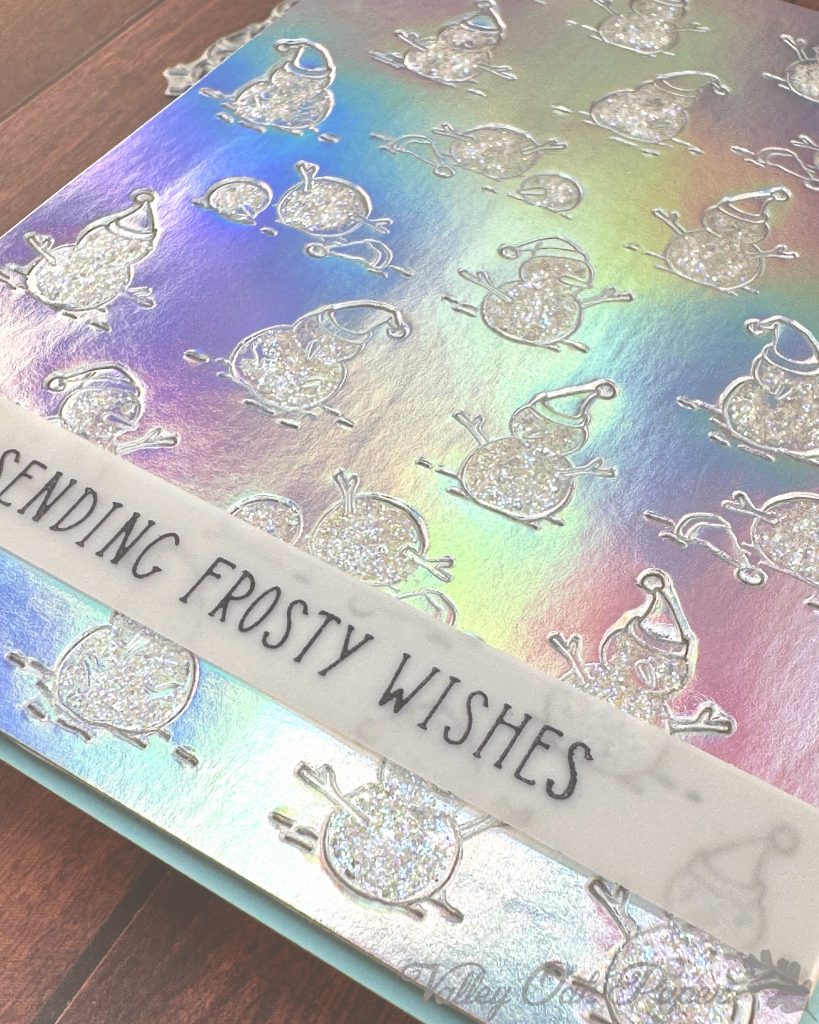 Closeup of a handmade greeting card on a wooden surface. The background on the card is made of silver holographic cardstock. It's been embossed with a pattern of snow people. The snow is covered with iridescent glitter.
Toward the bottom of the card, a thin vellum strip reads "Sending Frosty Wishes."
A slim matte of pale blue is visible around the patterned holographic cardstock.
Peeking out from under the card are some BetterPress plates. Some white flowers are also visible.