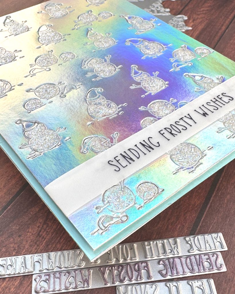 Closeup of a handmade greeting card on a wooden surface. The background on the card is made of silver holographic cardstock. It's been embossed with a pattern of snow people. The snow is covered with iridescent glitter.
Toward the bottom of the card, a thin vellum strip reads "Sending Frosty Wishes."
A slim matte of pale blue is visible around the patterned holographic cardstock.
Peeking out from under the card are some BetterPress plates.