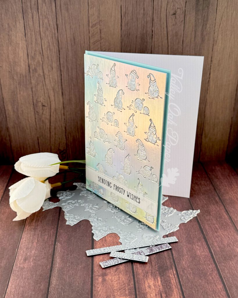 A handmade greeting card stands facing left on a wooden surface. The background on the card is made of silver holographic cardstock. It's been embossed with a pattern of snow people. The snow is covered with iridescent glitter.
Toward the bottom of the card, a thin vellum strip reads "Sending Frosty Wishes."
A slim matte of pale blue is visible around the patterned holographic cardstock.
Peeking out from under the card are some BetterPress plates. Some white flowers are also visible in the background.