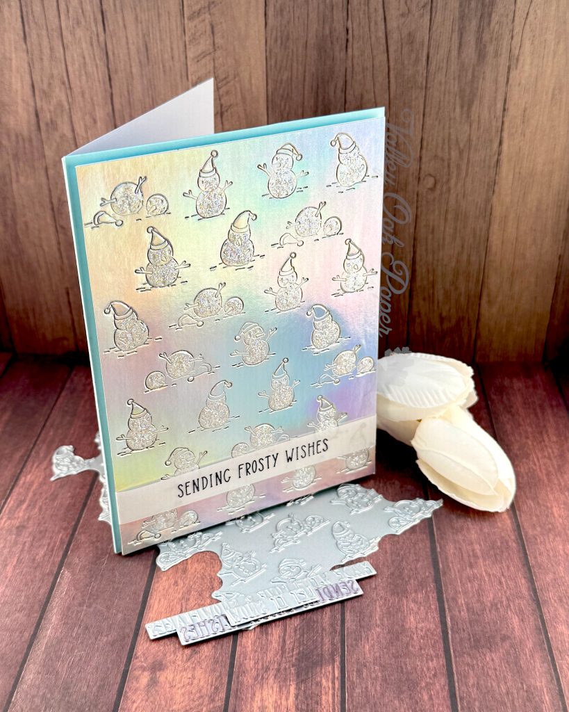 A handmade greeting card stands facing right on a wooden surface. The background on the card is made of silver holographic cardstock. It's been embossed with a pattern of snow people. The snow is covered with iridescent glitter.
Toward the bottom of the card, a thin vellum strip reads "Sending Frosty Wishes."
A slim matte of pale blue is visible around the patterned holographic cardstock.
Peeking out from under the card are some BetterPress plates. Some white flowers are also visible.