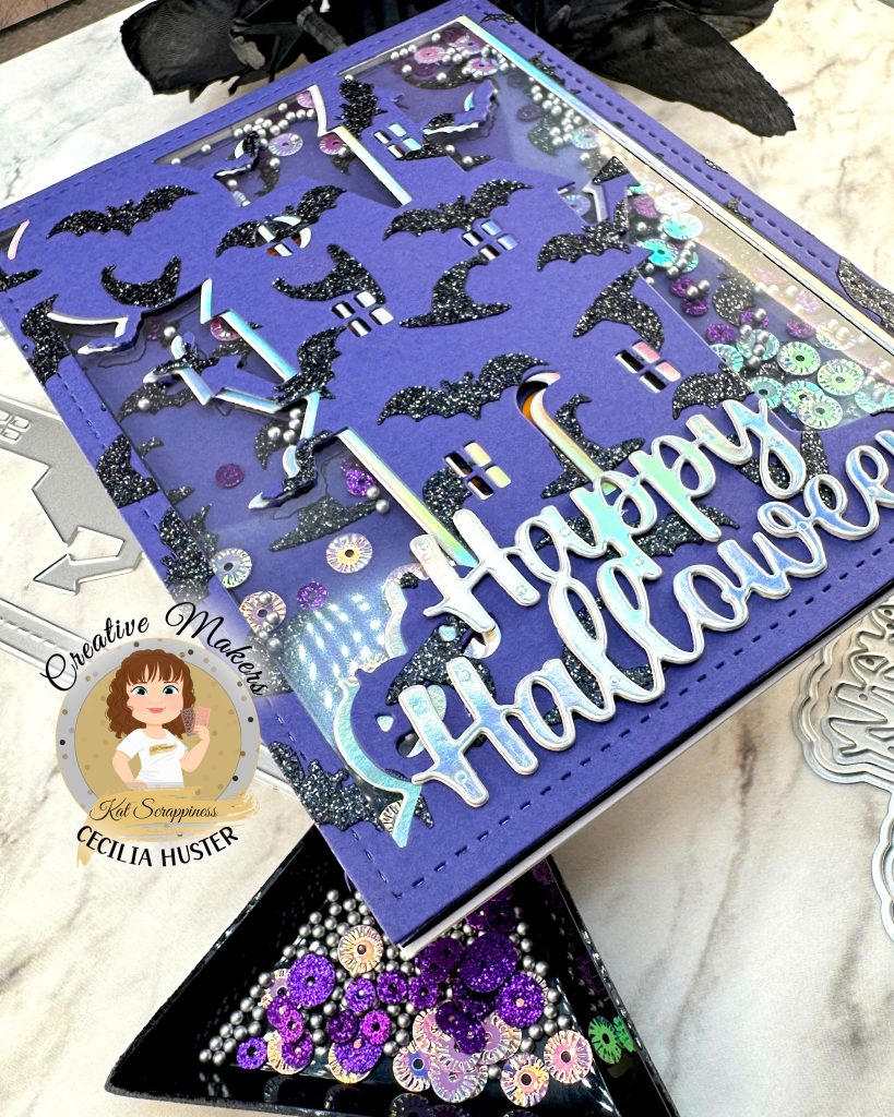 A close-up of a shaker greeting card in purple, black and silver is pictured against a white marble background. The card has an overall pattern of black glitter witchy hats and bats. The motif is a haunted house. Against the background we can see sequins in purple and silver tones. The silver sentiment reads Happy Halloween. This is a close-up of the lower left corner. 

The cover die, sentiment die and sequins that were used to create the card are also displayed. The little plastic triangle tray with the sequins is in focus. In addition, some black roses can be seen. In the lower left corner is a watermark that reads Creative Maker Cecilia Huster for Kat Scrappiness. 