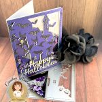 A shaker greeting card in purple, black and silver is pictured against a warm wood background. The card has an overall pattern of black glitter witchy hats and bats. The motif is a haunted house. Against the background we can see sequins in purple and silver tones. The silver sentiment reads Happy Halloween. The card faces right. The cover die, sentiment die and sequins that were used to create the card are also displayed. In addition, some black roses can be seen. In the lower left corner is a watermark that reads Creative Maker Cecilia Huster for Kat Scrappiness.