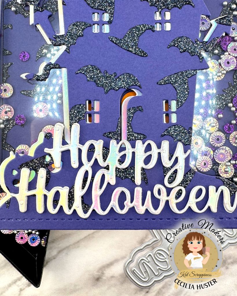 A close-up of a shaker greeting card in purple, black and silver is pictured against a white marble background. The card has an overall pattern of black glitter witchy hats and bats. The motif is a haunted house. Against the background we can see sequins in purple and silver tones. The silver sentiment reads Happy Halloween. This is a close-up of the sentiment. 

The cover die, sentiment die and sequins that were used to create the card are also displayed. In addition, some black roses can be seen. In the lower right corner is a watermark that reads Creative Maker Cecilia Huster for Kat Scrappiness. 