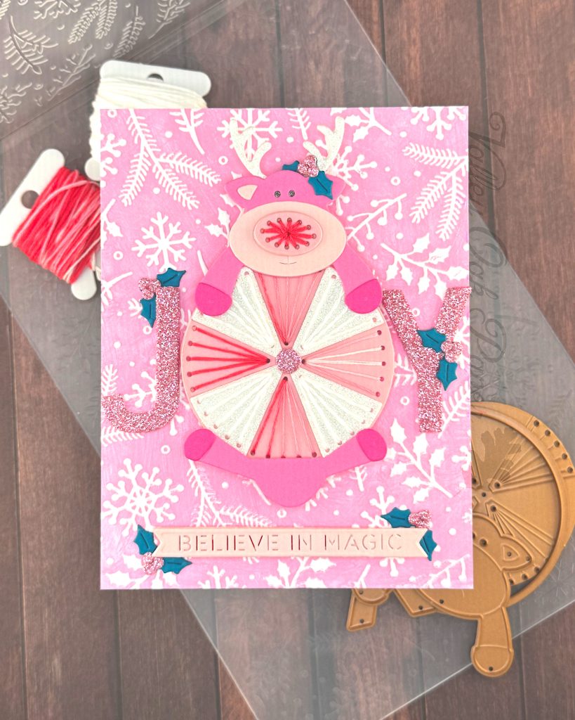 Handmade greeting card featuring a pink reindeer and an oversized peppermint candy against a pink winter-themed background with white snowflakes and pine branches.
The reindeer has white glitter antlers accented with small teal holly leaves, pink hooves, and an embroidered nose.
The peppermint candy has been stitched in alternating white and variegated pink embroidery floss.
The main sentiment on the card, JOY, is formed by the letters J and Y on either side of the round candy. The text 'BELIEVE IN MAGIC' appears near the bottom.
The card is displayed on a wooden surface alongside crafting supplies including, an embossing folder, embroidery floss and dies.