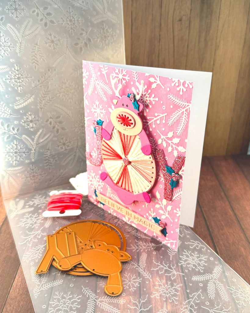 Handmade greeting card featuring a pink reindeer and an oversized peppermint candy against a pink winter-themed background with white snowflakes, holly and pine branches. The card is standing up facing to the left.
The reindeer has white glitter antlers accented with small teal holly leaves, pink hooves, and an embroidered nose.
The peppermint candy has been stitched in alternating white and variegated pink embroidery floss.
The main sentiment on the card, JOY, is formed by the letters J and Y on either side of the round candy. The text 'BELIEVE IN MAGIC' appears near the bottom.
The card is displayed on a wooden surface alongside crafting supplies including, an embossing folder, embroidery floss and dies."