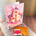 "Handmade greeting card featuring a pink reindeer and an oversized peppermint candy against a pink winter-themed background with white snowflakes and pine branches. The reindeer has white glitter antlers accented with small teal holly leaves, pink hooves, and an embroidered nose. The peppermint candy has been stitched in alternating white and variegated pink embroidery floss. The main sentiment on the card, JOY, is formed by the letters J and Y on either side of the round candy. The text 'BELIEVE IN MAGIC' appears near the bottom. The card is displayed on a wooden surface alongside crafting supplies including, an embossing folder, embroidery floss and dies."