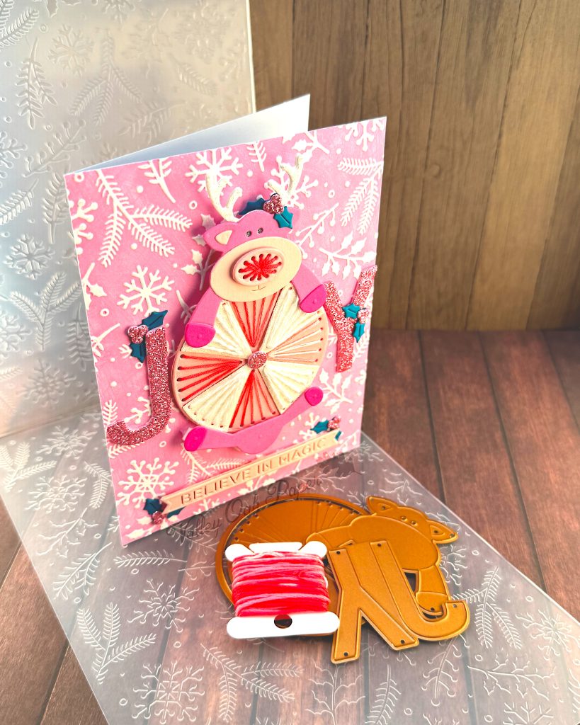 Handmade greeting card featuring a pink reindeer and an oversized peppermint candy against a pink winter-themed background with white snowflakes, holly and pine branches. The card stands up facing right.
The reindeer has white glitter antlers accented with small teal holly leaves, pink hooves, and an embroidered nose.
The peppermint candy has been stitched in alternating white and variegated pink embroidery floss.
The main sentiment on the card, JOY, is formed by the letters J and Y on either side of the round candy. The text 'BELIEVE IN MAGIC' appears near the bottom.
The card is displayed on a wooden surface alongside crafting supplies including, an embossing folder, embroidery floss and dies.