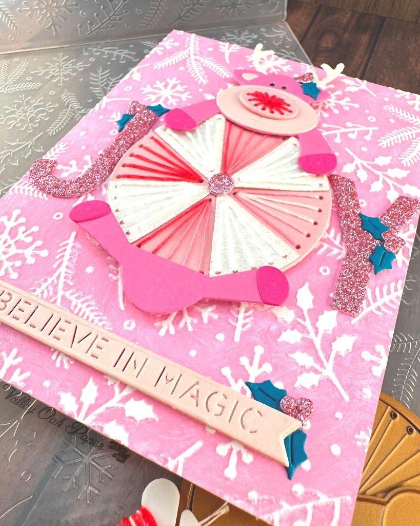 Handmade greeting card featuring a pink reindeer and an oversized peppermint candy against a pink winter-themed background with white snowflakes and pine branches.
The reindeer has white glitter antlers accented with small teal holly leaves, pink hooves, and an embroidered nose.
The peppermint candy has been stitched in alternating white and variegated pink embroidery floss.
The main sentiment on the card, JOY, is formed by the letters J and Y on either side of the round candy. The text 'BELIEVE IN MAGIC' appears near the bottom.
The card is displayed on a wooden surface alongside crafting supplies including, an embossing folder and dies.