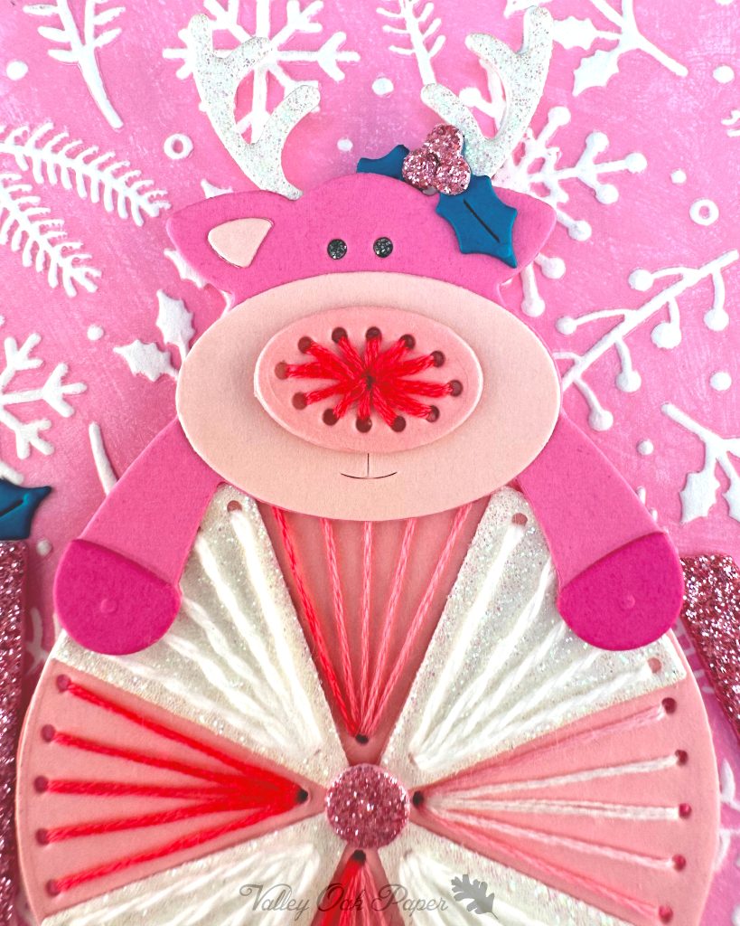 Closeup of a handmade greeting card featuring a pink reindeer and an oversized peppermint candy against a pink winter-themed background with white snowflakes and pine branches.
The reindeer has white glitter antlers accented with small teal holly leaves, pink hooves, and an embroidered nose.
The peppermint candy has been stitched in alternating white and variegated pink embroidery floss.