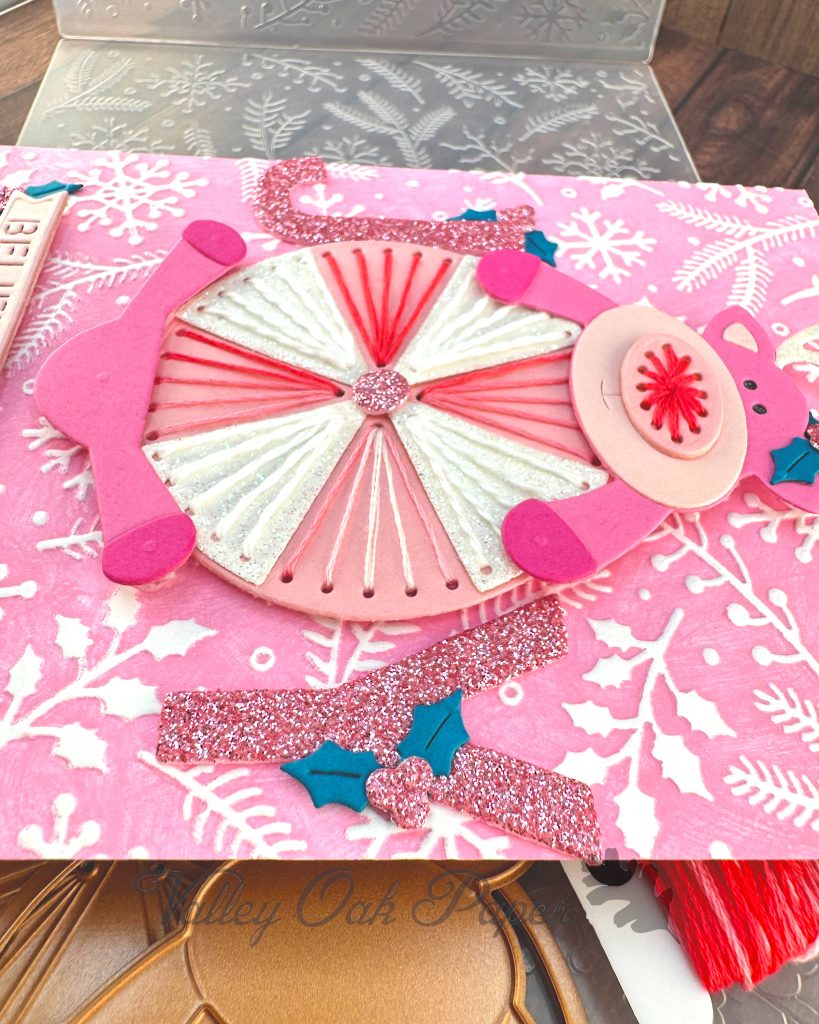 Handmade greeting card seen from the side. The card features a pink reindeer and an oversized peppermint candy against a pink winter-themed background with white snowflakes and pine branches.
The reindeer has pink hooves, and an embroidered nose.
The peppermint candy has been stitched in alternating white and variegated pink embroidery floss.
The main sentiment on the card, JOY, is formed by the letters J and Y on either side of the round candy.
The card is displayed on a wooden surface alongside crafting supplies including, an embossing folder, embroidery floss and dies.