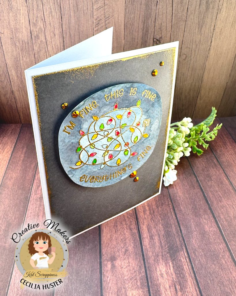 A handmade greeting card with a grey background outlined in grungy gold. The main feature of the card is a watercolor panel showing a tangled light string with lights in red, amber and green.
The text around the image says "I'm fine. This is fine. Everything's fine." A few gold gems adorn the card front.
It's photographed on a rustic wooden background. In the lower left corner is a watermark with the text "Creative Maker for Kat Scrappiness: Cecilia Huster."