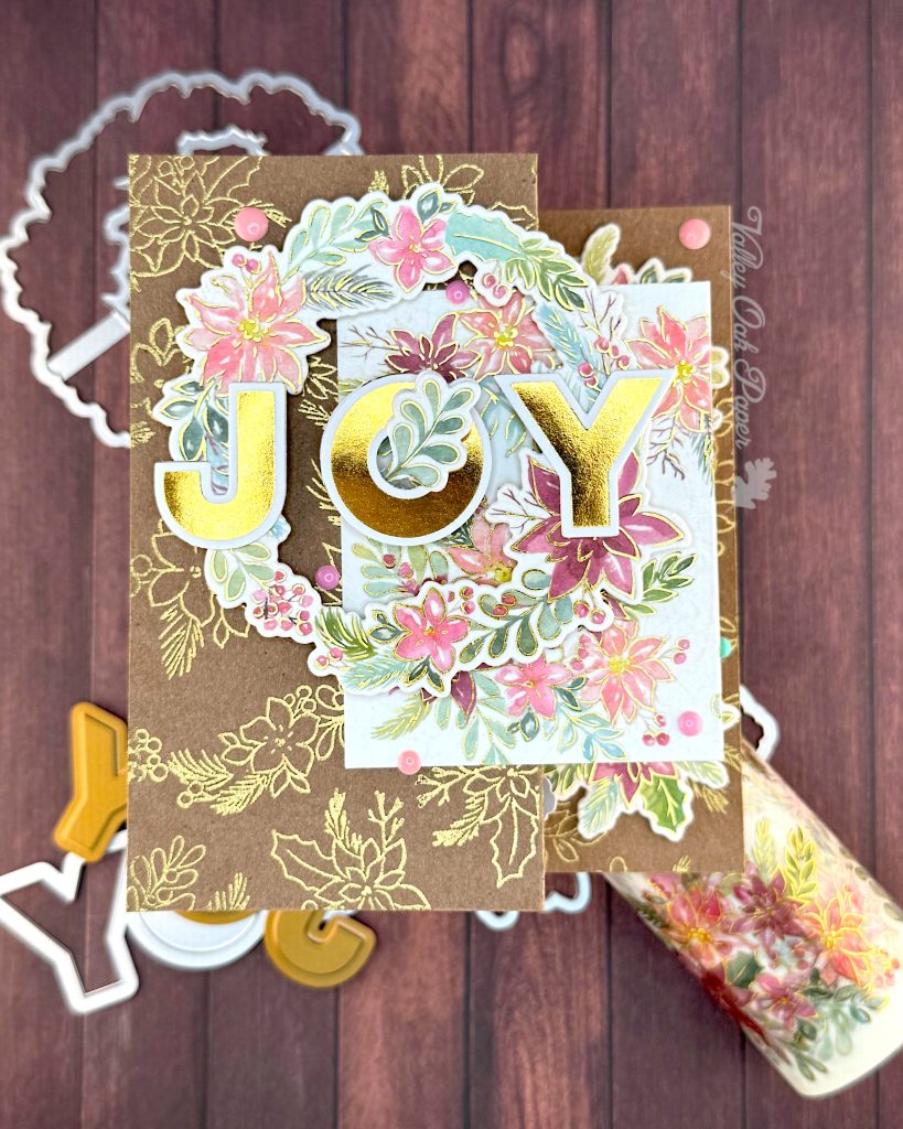 Handmade double Z-fold greeting card featuring the word 'JOY' in gold metallic letters centered on layered panels.
The card displays an elaborate floral wreath arrangement with pink, coral, and burgundy watercolor holiday flowers surrounded by sage green and teal leaves. Gold heat-embossed botanical designs embellish the kraft paper background layers.
The card is photographed on a rustic wood surface with dies and foil plates visible at the edges and a matching holiday floral washi tape roll visible in the lower right corner.