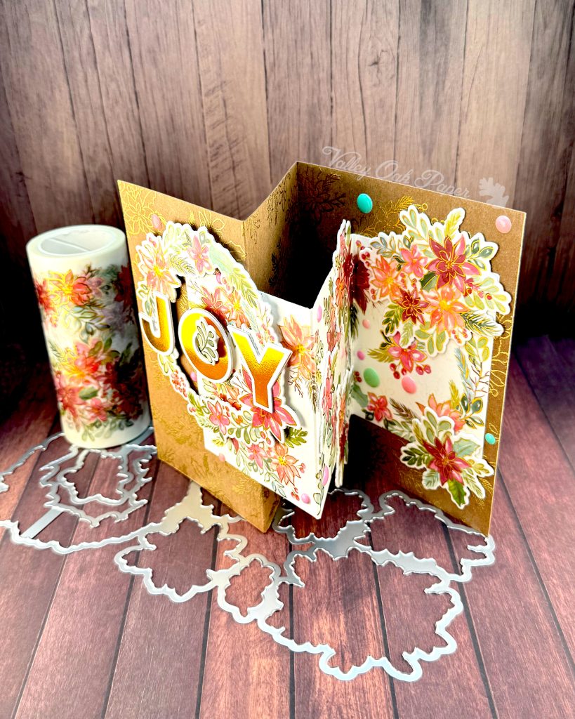 Handmade double Z-fold greeting card featuring the word 'JOY' in gold metallic letters centered on layered panels of Kraft cardstock and cream cardstock.
The card displays elaborate floral arrangements with pink, coral, and burgundy watercolor holiday flowers surrounded by sage green and teal leaves. Gold heat-embossed botanical designs embellish the kraft paper background layers.
The card is photographed standing on a rustic wood surface with dies and foil plates and a matching holiday floral washi tape roll visible behind it.