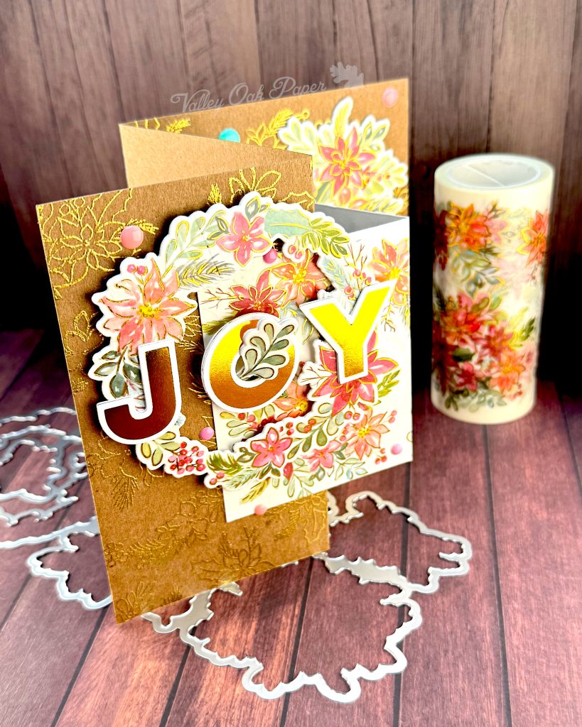 Handmade double Z-fold greeting card featuring the word 'JOY' in gold metallic letters centered on layered panels of Kraft cardstock and cream cardstock.
The card displays elaborate floral arrangements with pink, coral, and burgundy watercolor holiday flowers surrounded by sage green and teal leaves. Gold heat-embossed botanical designs embellish the kraft paper background layers.
The card is photographed standing on a rustic wood surface with dies and foil plates and a matching holiday floral washi tape roll visible behind it.