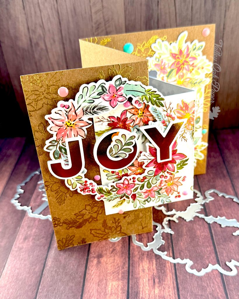 Handmade double Z-fold greeting card featuring the word 'JOY' in gold metallic letters centered on layered panels of Kraft cardstock and cream cardstock.
The card displays elaborate floral arrangements with pink, coral, and burgundy watercolor holiday flowers surrounded by sage green and teal leaves. Gold heat-embossed botanical designs embellish the kraft paper background layers.
The card is photographed standing on a rustic wood surface with dies.