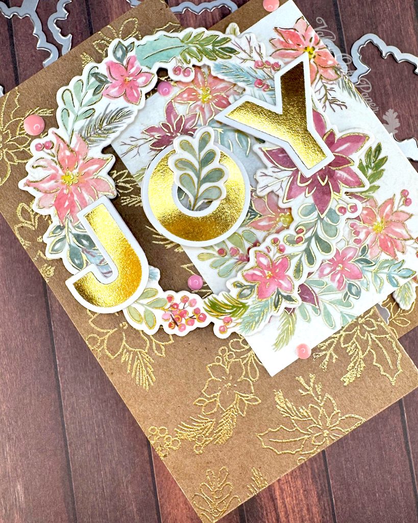 Handmade double Z-fold greeting card featuring the word 'JOY' in gold metallic letters centered on layered panels.
The card displays an elaborate floral wreath arrangement with pink, coral, and burgundy watercolor holiday flowers surrounded by sage green and teal leaves. Gold heat-embossed botanical designs embellish the kraft paper background layers.
The card is photographed on a rustic wood surface with dies and foil plates visible at the edges.