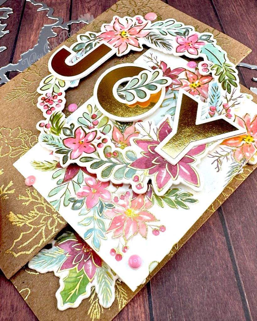 Handmade double Z-fold greeting card featuring the word 'JOY' in gold metallic letters centered on layered panels.
The card displays an elaborate floral wreath arrangement with pink, coral, and burgundy watercolor holiday flowers surrounded by sage green and teal leaves. Gold heat-embossed botanical designs embellish the kraft paper background layers.
The card is photographed on a rustic wood surface with dies and foil plates visible at the edges.