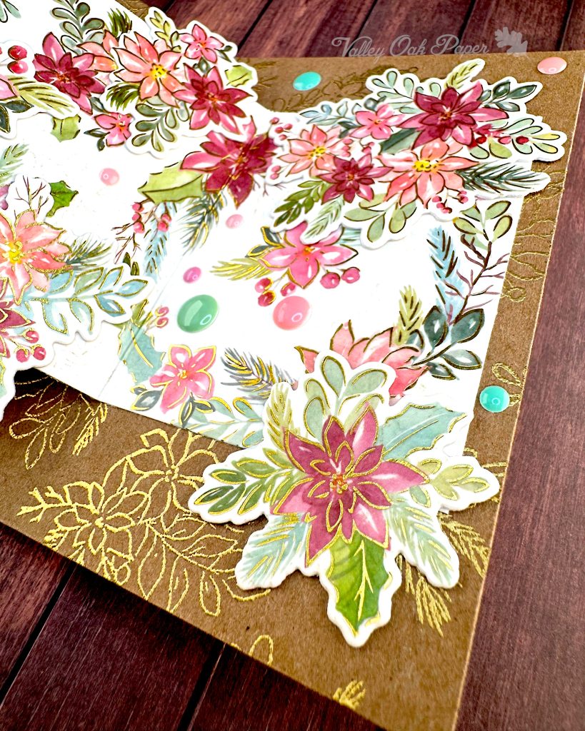 Closeup of the last panel of a handmade double Z-fold greeting card with layered panels of Kraft cardstock and cream cardstock.
The card displays elaborate floral arrangements with pink, coral, and burgundy watercolor holiday flowers surrounded by sage green and teal leaves on the smaller panel. Gold heat-embossed botanical designs embellish the kraft paper background layers.
Matching pink and green enamel dots have been scattered around the card.
The card is photographed lying on a rustic wood surface.