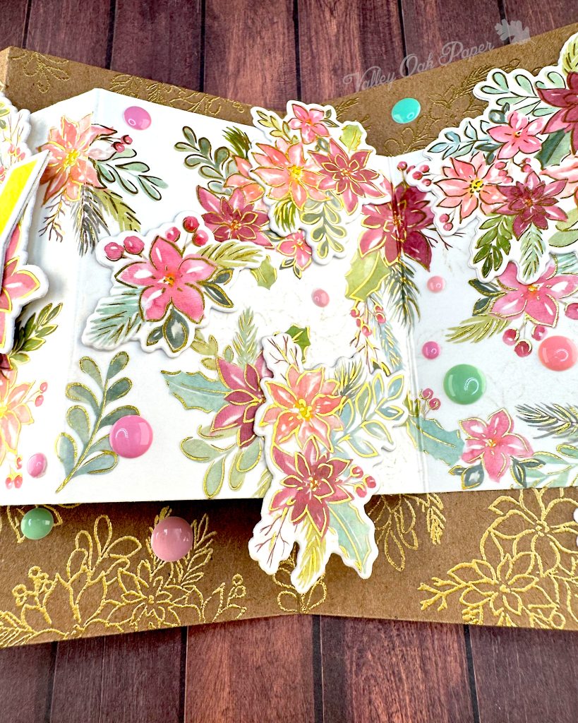 Closeup of the middle panel of a handmade double Z-fold greeting card with layered panels of Kraft cardstock and cream cardstock.
The card displays elaborate floral arrangements with pink, coral, and burgundy watercolor holiday flowers surrounded by sage green and teal leaves on the smaller panel. Gold heat-embossed botanical designs embellish the kraft paper background layers.
The card is photographed lying on a rustic wood surface.