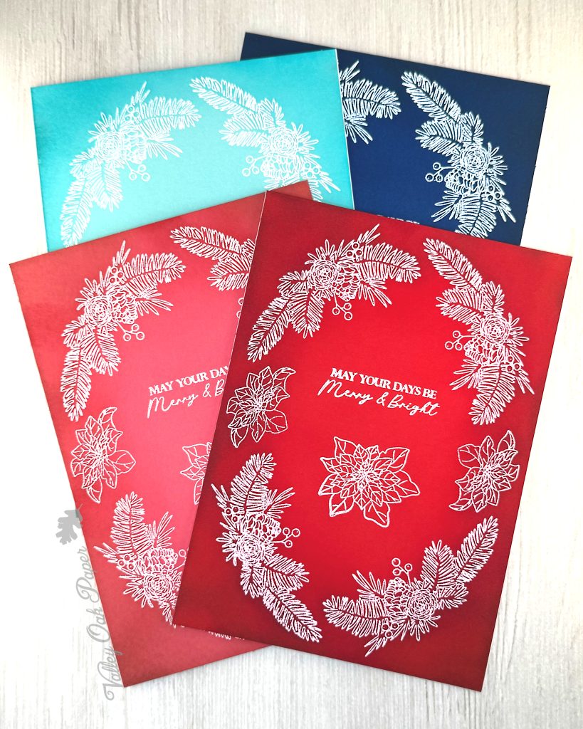 Four handmade greeting card that are 5" by 7" are lying on a white wooden background. They've all been heat-embossed in white with a floral holiday wreath arrangement. The cards are red, pink, navy and turquoise.
The sentiment reads "May Your Days be Merry & Bright." It sits in the center of the cards.
