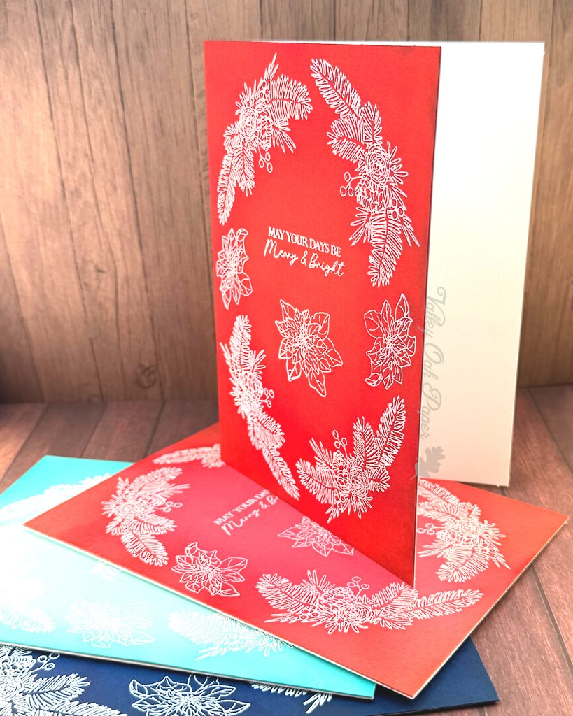 A handmade greeting card that is 5" by 7" stands on a small heap of similar cards. It is facing to the left. The cards have all been heat-embossed in white with a floral holiday wreath arrangement.
The sentiment reads "May Your Days be Merry & Bright." It sits in the center of the cards.
The cards have been photographed on a rustic wooden background.