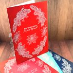 A handmade greeting card that is 5" by 7" stands on a small heap of similar cards. They've all been heat-embossed in white with a floral holiday wreath arrangement. The sentiment reads "May Your Days be Merry & Bright." It sits in the center of the cards. The cards have been photographed on a rustic wooden background.