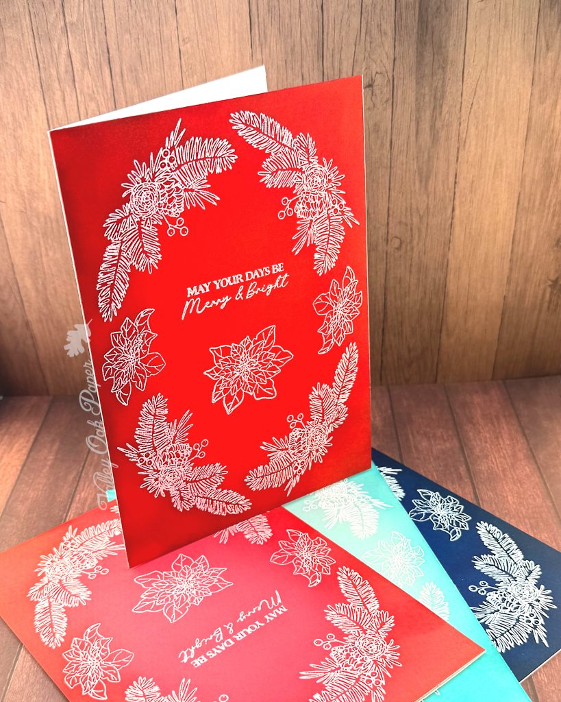 A handmade greeting card that is 5" by 7" stands on a small heap of similar cards. It is facing to the right. The cards have all been heat-embossed in white with a floral holiday wreath arrangement.
The sentiment reads "May Your Days be Merry & Bright." It sits in the center of the cards.
The cards have been photographed on a rustic wooden background.