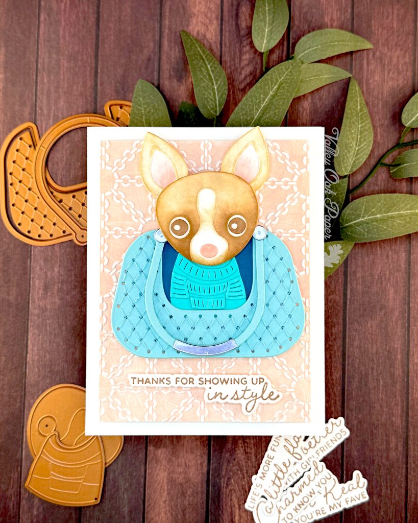 A handmade greeting card featuring a chihuahua wearing a turquoise sweater, sitting in a matching quilted bag. The bag is centered on a peach background embossed with a white chain pattern. The greeting reads 'THANKS FOR SHOWING UP in style' at the bottom.
The card is photographed on a dark wood surface alongside paper craft elements including dog-shaped dies and sentiments, with green sage leaves in the background.