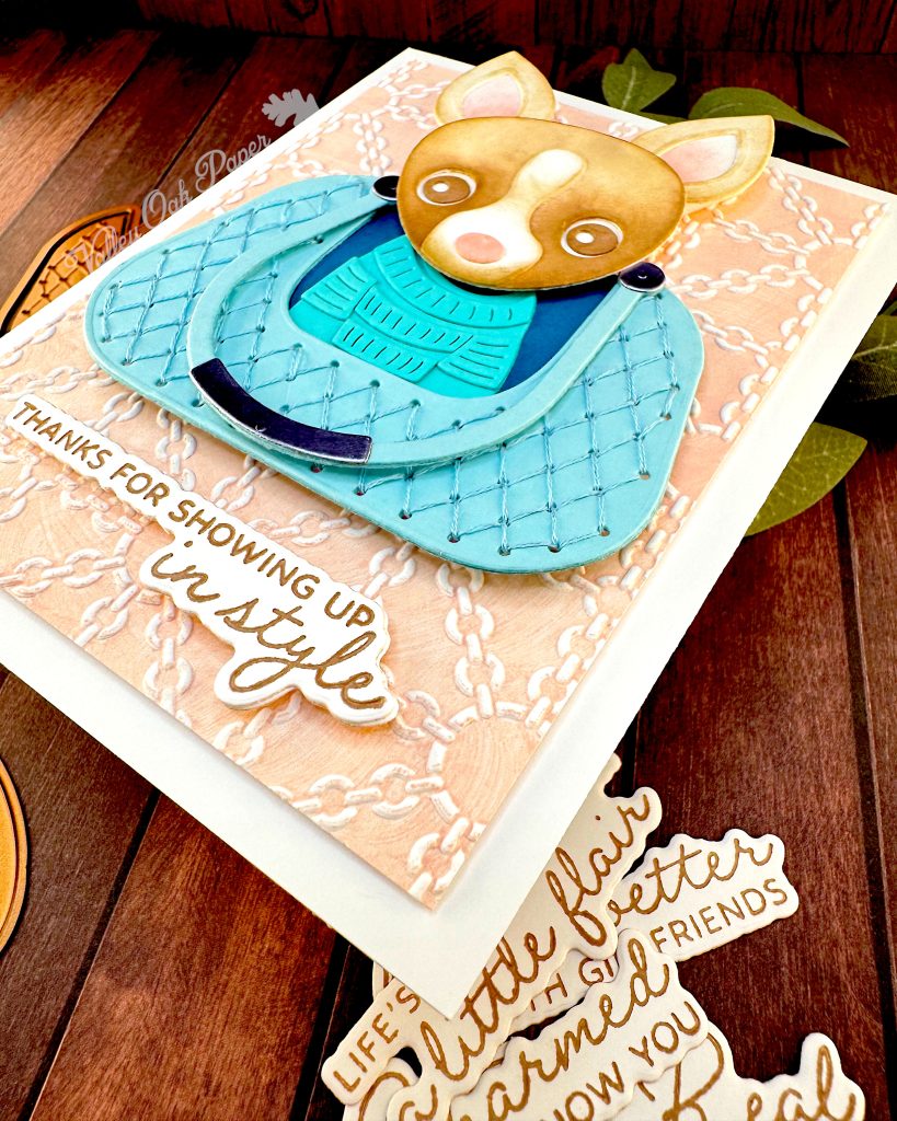 A handmade greeting card featuring a chihuahua wearing a turquoise sweater, sitting in a matching quilted bag. The bag is centered on a peach background embossed with a white chain pattern. The greeting reads 'THANKS FOR SHOWING UP in style' at the bottom.
The angle of this photo highlights the texture of the peach embossed background.
The card is photographed on a dark wood surface alongside paper craft elements including dog-shaped dies and sentiments, with green sage leaves in the background.