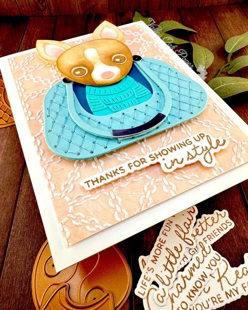 A handmade greeting card featuring a chihuahua wearing a turquoise sweater, sitting in a matching quilted bag. The bag is centered on a peach background embossed with a white chain pattern. The greeting reads 'THANKS FOR SHOWING UP in style' at the bottom.
The angle of this photo highlights the texture of the peach embossed background and the elevated sentiment.
The card is photographed on a dark wood surface alongside paper craft elements including dog-shaped dies and sentiments, with green sage leaves in the background.