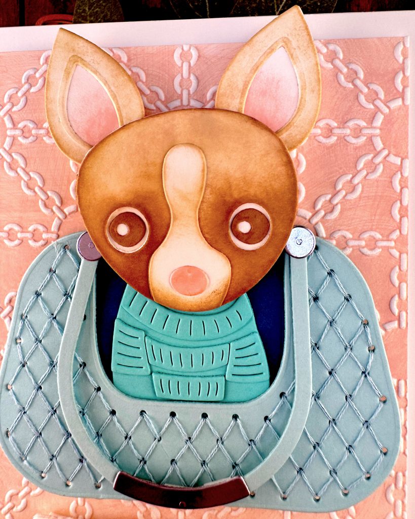 A closeup of a handmade greeting card featuring a chihuahua wearing a turquoise sweater, sitting in a matching quilted bag. The bag is centered on a peach background embossed with a white chain pattern. The chihuahua is brown with a white blaze. Its nose is pink. It looks anxious.
The card is photographed on a dark wood surface alongside paper craft elements including dog-shaped dies and sentiments, with green sage leaves in the background.