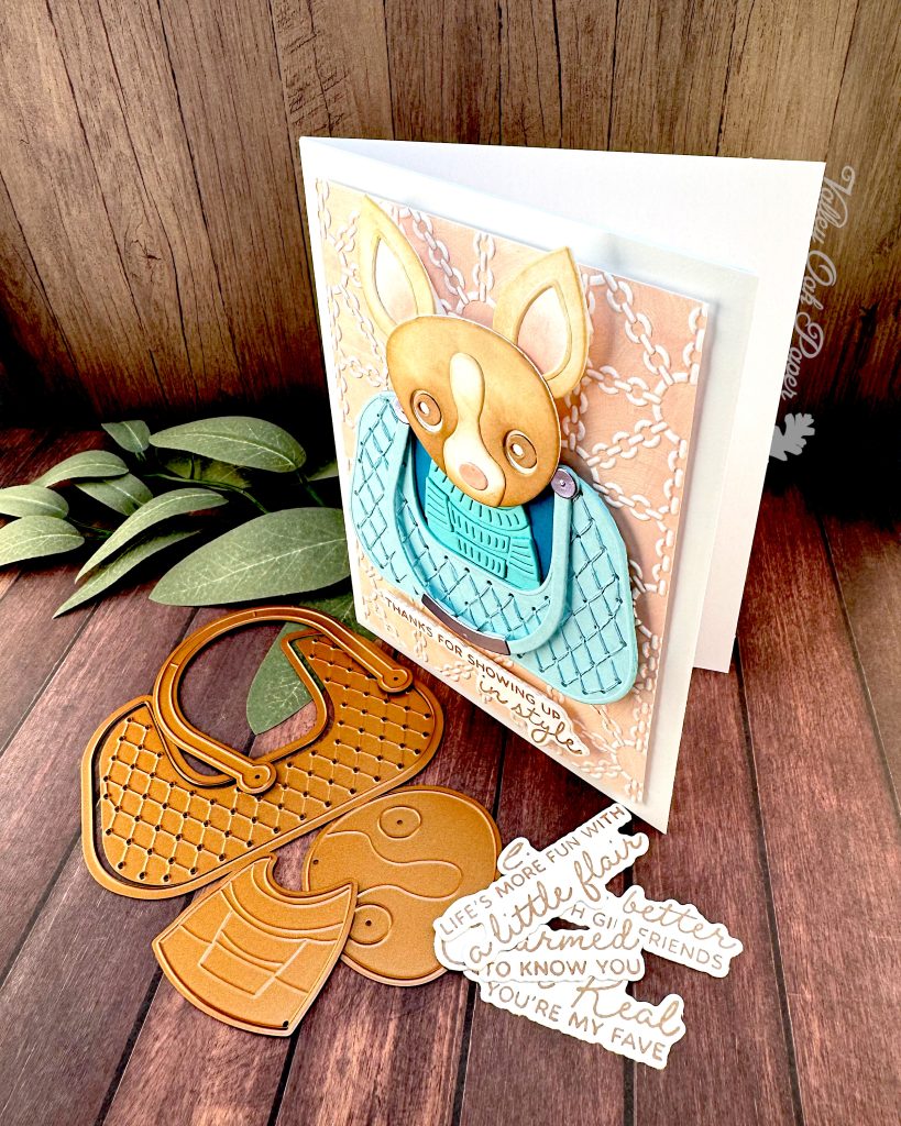 A handmade greeting card featuring a chihuahua wearing a turquoise sweater, sitting in a matching quilted bag. The bag is centered on a peach background embossed with a white chain pattern. The greeting reads 'THANKS FOR SHOWING UP in style' at the bottom.
The card stands facingleft on a dark wood surface alongside paper craft elements including dog-shaped dies and sentiments, with green sage leaves in the background.