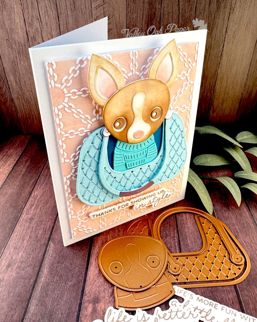 A handmade greeting card featuring a chihuahua wearing a turquoise sweater, sitting in a matching quilted bag. The bag is centered on a peach background embossed with a white chain pattern. The greeting reads 'THANKS FOR SHOWING UP in style' at the bottom.
The card stands facing right on a dark wood surface alongside paper craft elements including dog-shaped dies and sentiments, with green sage leaves in the background.