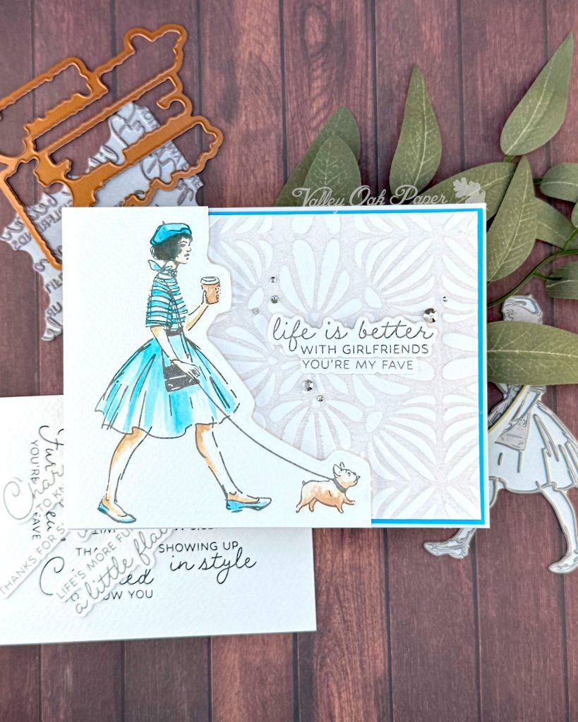 A handmade greeting card featuring an illustration of a fashionable woman in a turquoise striped shirt, fluffy turquoise skirt, and matching beret walking a small fat dog while holding a coffee cup.
The background is embossed with an organic, floral pattern.
The sentiment reads'Life is better with girlfriends. You're my fave' and is surrounded by small silver rhinestone embellishments.
The card is matted with a bright turquoise border. It sits on a rustic wooden background alongside crafting supplies such as BetterPress plates and dies.